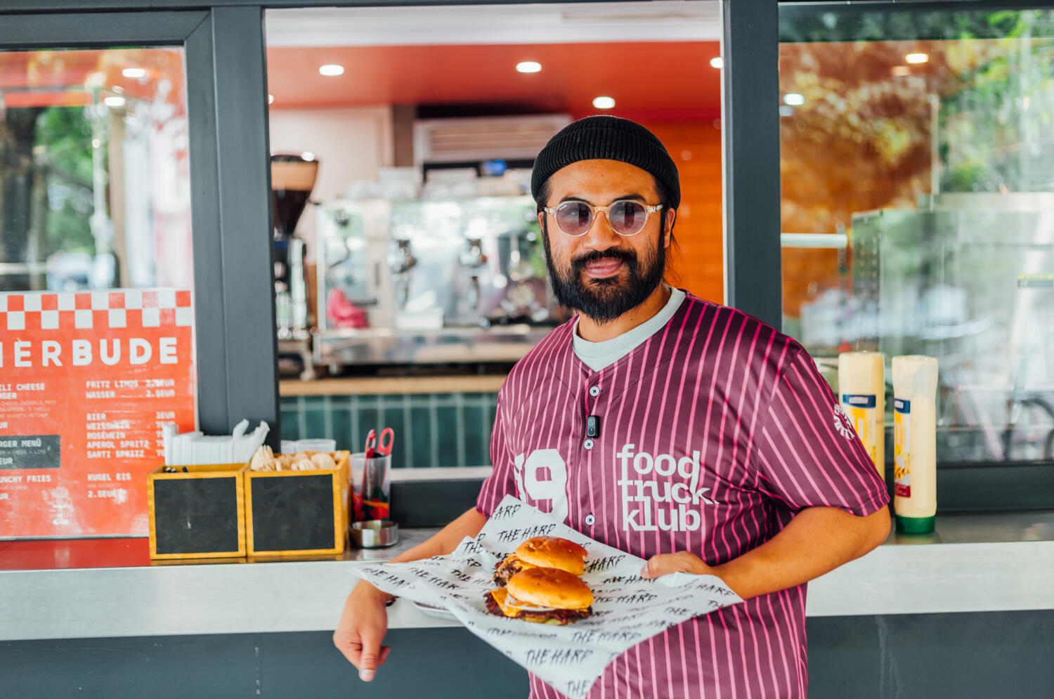 Chef Nucki der Probierbude Hannover steht vor dem Tresen und präsentiert zwei frisch zubereitete Burger auf Papier.